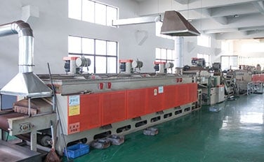 Büyük pencereleri olan bir fabrikanın yeşil zeminlerinde kırmızı ve gri endüstriyel makineler sıra halinde duruyor.