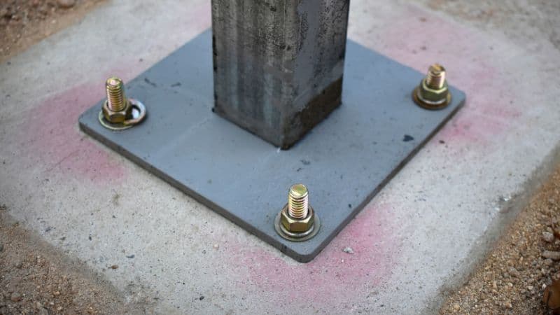 A metal post base plate is bolted to concrete with four anchors, using wedge or sleeve types for secure installation.