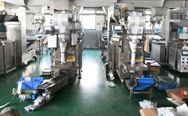 Máquinas de embalagem em uma fábrica com sacos, materiais no chão e equipamentos ao redor da área de trabalho.